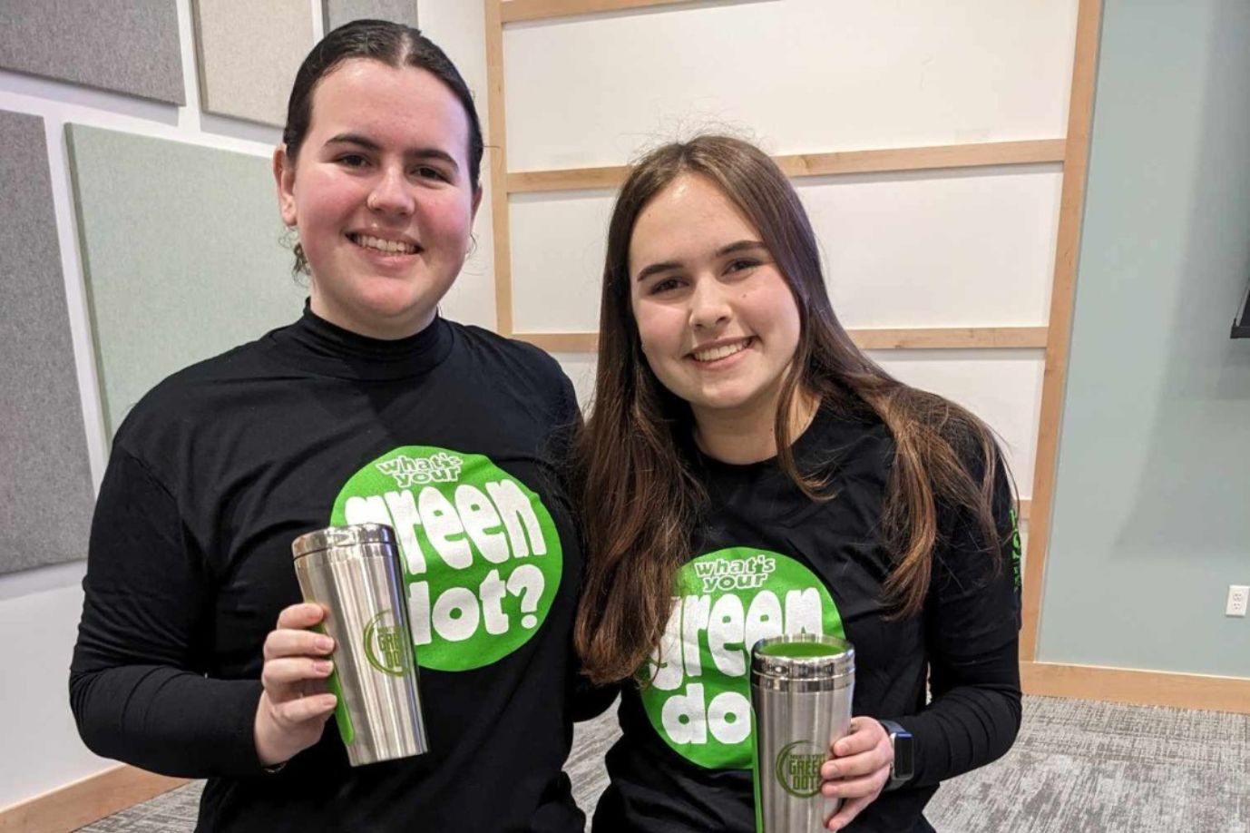 Two students pose for a photo wearing Green Dot Tees while holding a tumblers that have the Green Dot logo on them