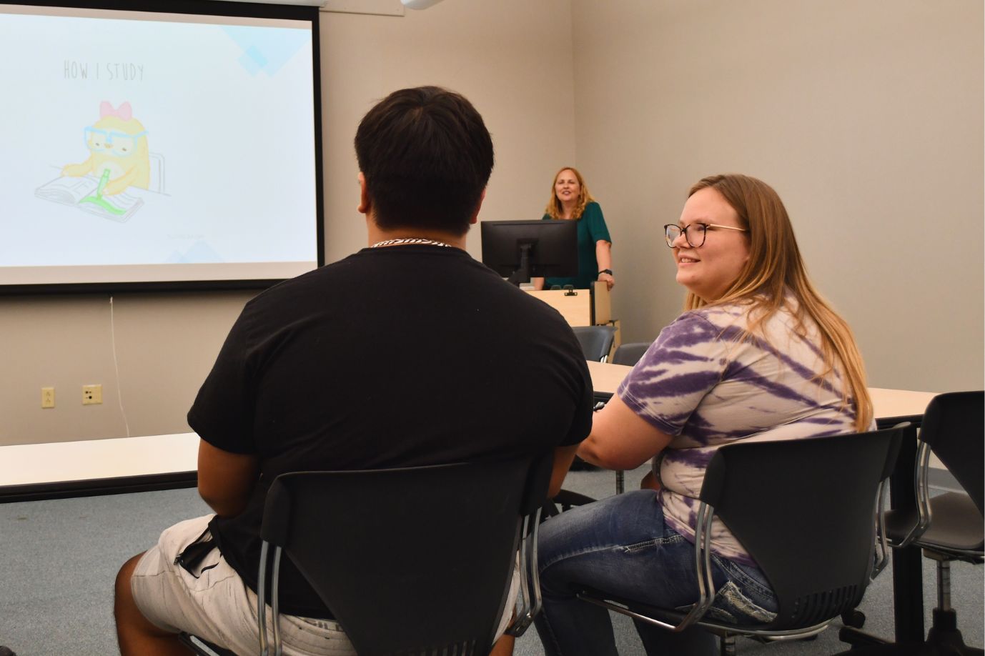 Two students sitting in a workshop about study skills while a presenter speaks from a podium. The PowerPoint slide reads "How do you study?"