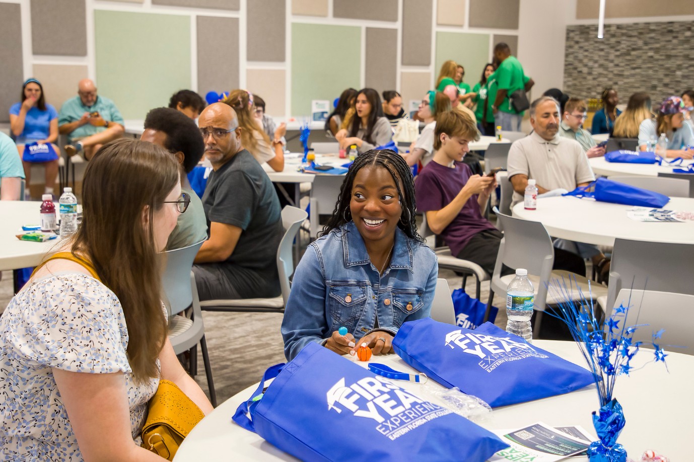 Students sit in a large conference room at round tables. They smile as they speak to each other. There are blue tote bags on the tables that says First Year Experience