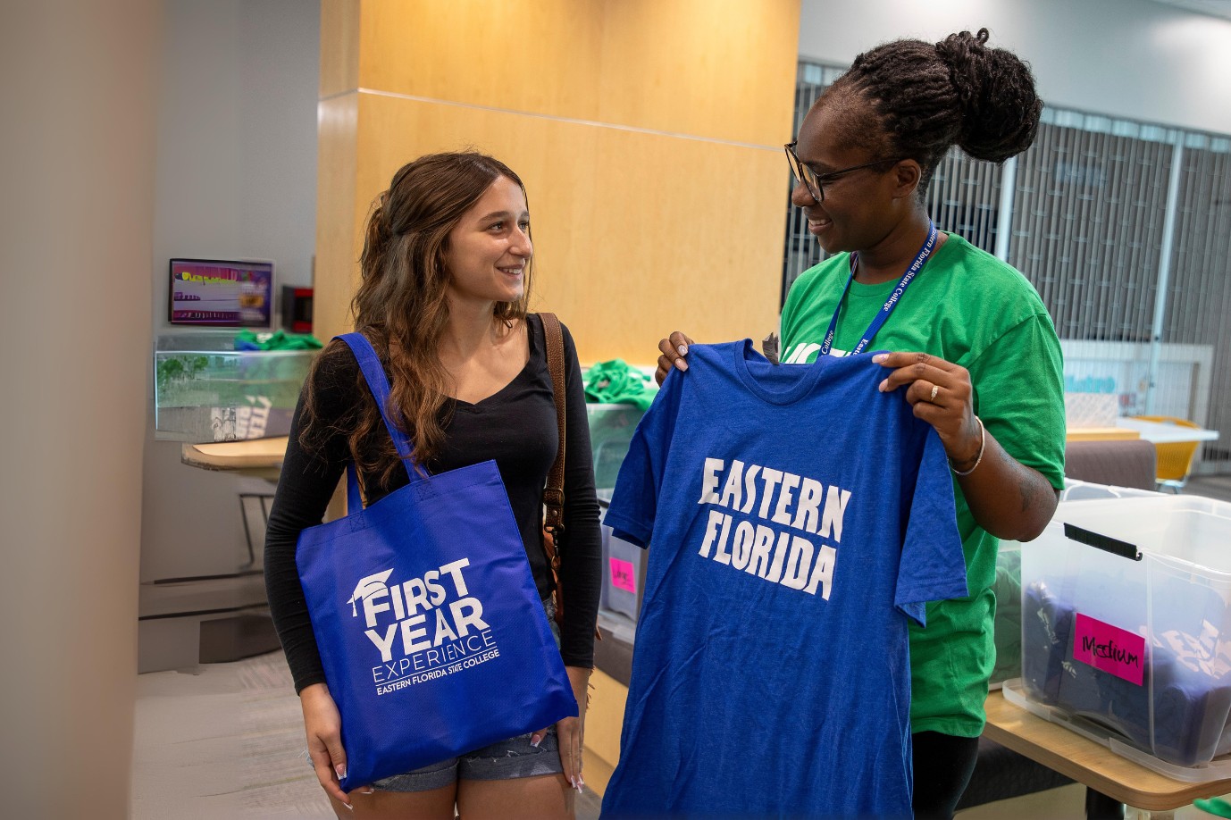 An EFSC staff member presents a student with an EFSC branded shirt. The female student is wearing a blue tote bag that says First Year Experience.