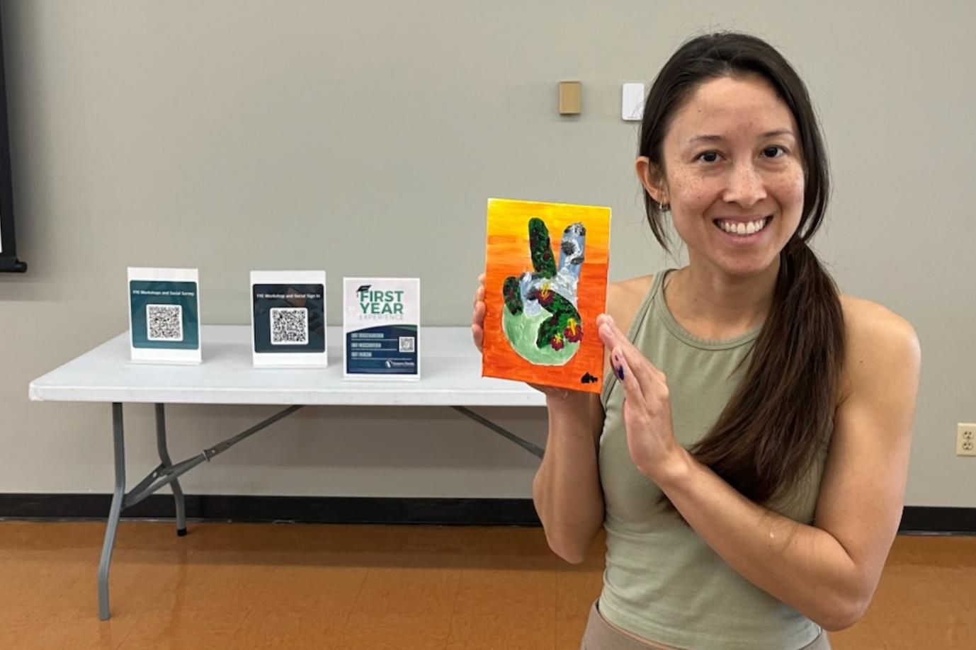 A smiling student is holding up a painting that they made during a First Year Experience event.